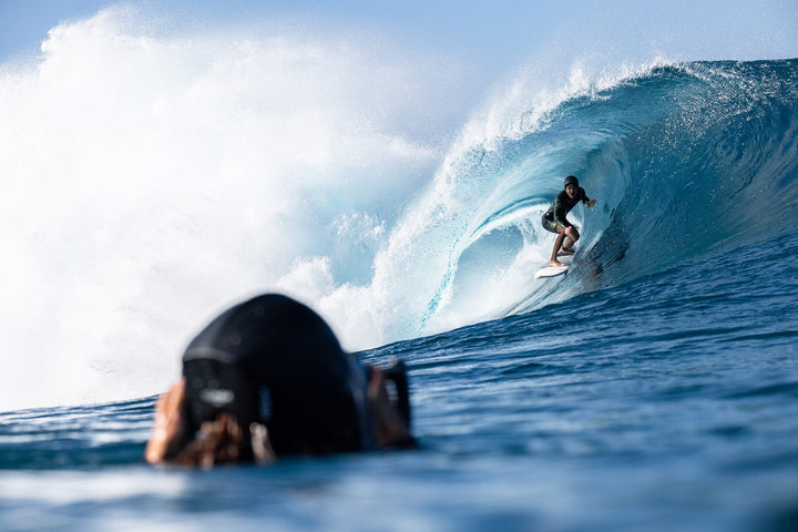 A wetsuit-clad surfer wearing a hood crouches inside a huge barreling wave while a water photographer in a black hood captures the action from the water in the foreground.
