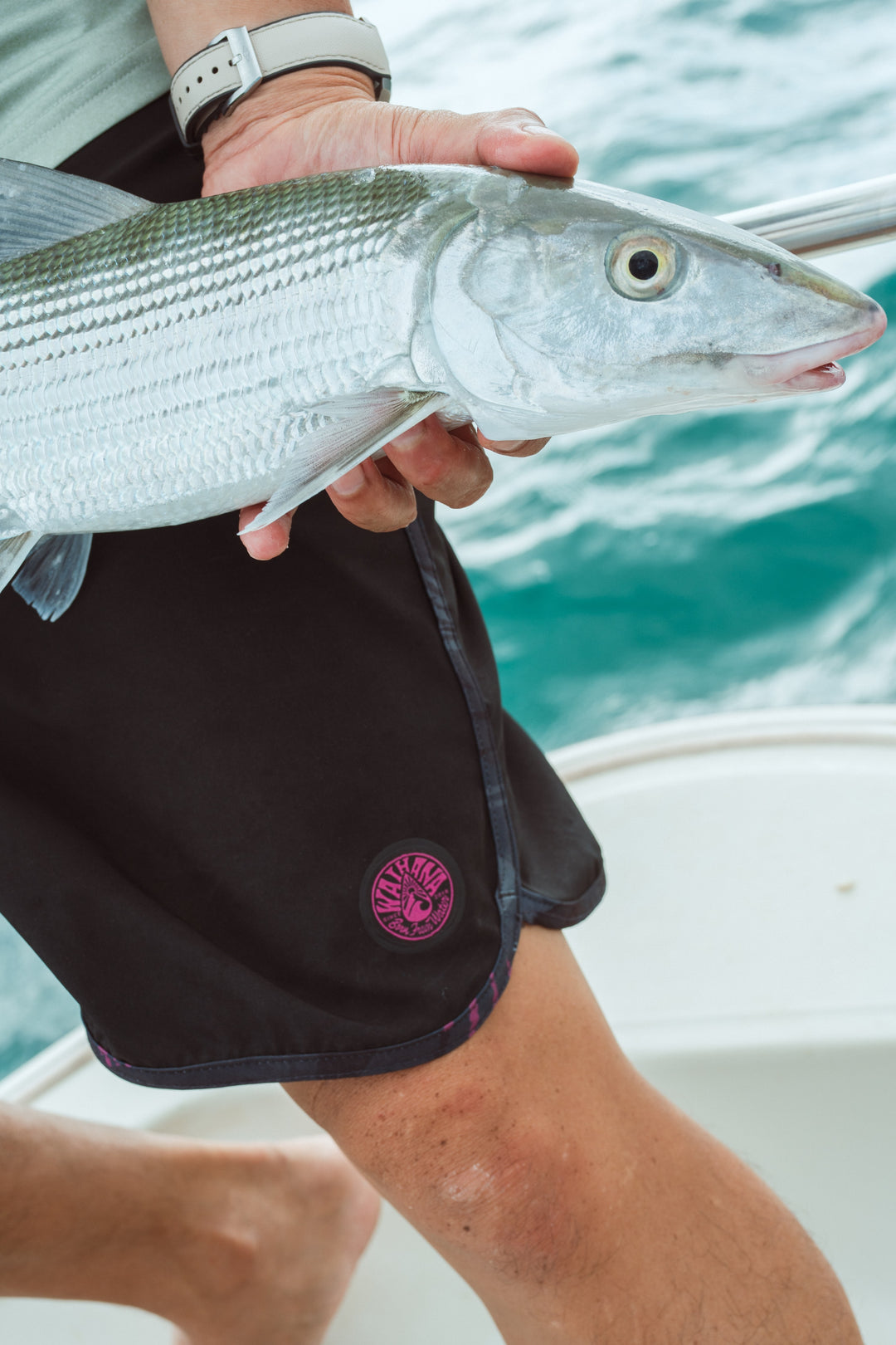 Person in black Waihana board shorts with pink logo holding a silver bonefish on a boat over turquoise water in Kaneohe Bay.