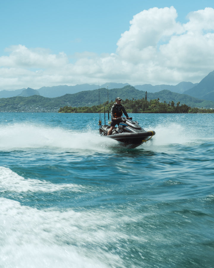 A man in a rashguard and hat rides a fishing-equipped jet ski at high speed across the blue waters of Kaneohe Bay, Hawaii, with tropical mountains visible in the background.
