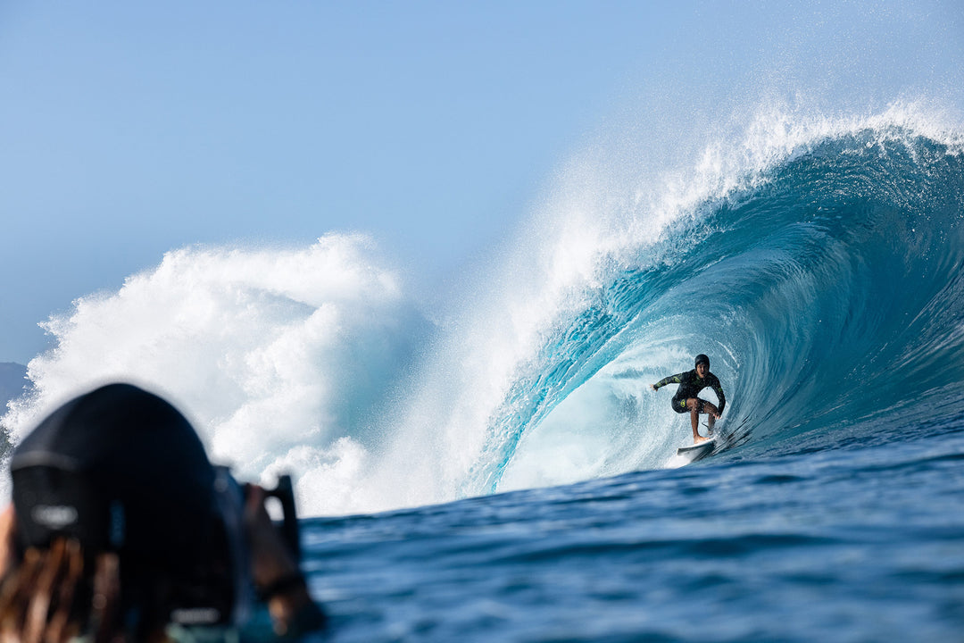 A wetsuit-clad surfer crouches inside a massive turquoise barrel wave while a water photographer shoots from the foreground on a bright blue-sky day.
