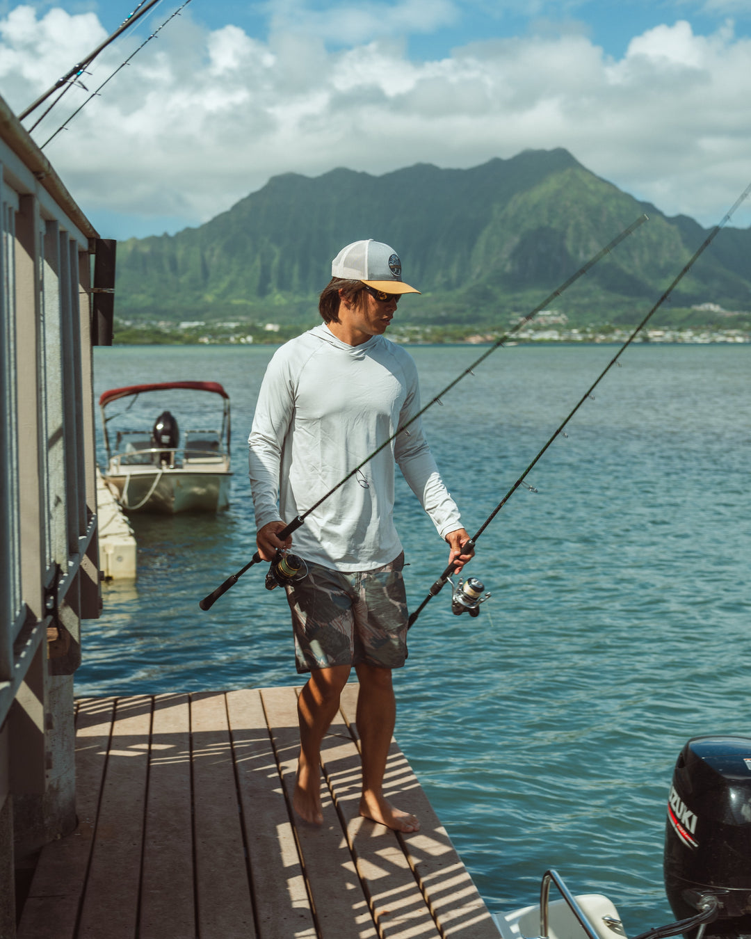 A man in a white long-sleeve rashguard and snapback hat carries two fishing rods along a wooden dock at Kaneohe Bay, Hawaii, with a moored boat and dramatic green mountains visible behind him.