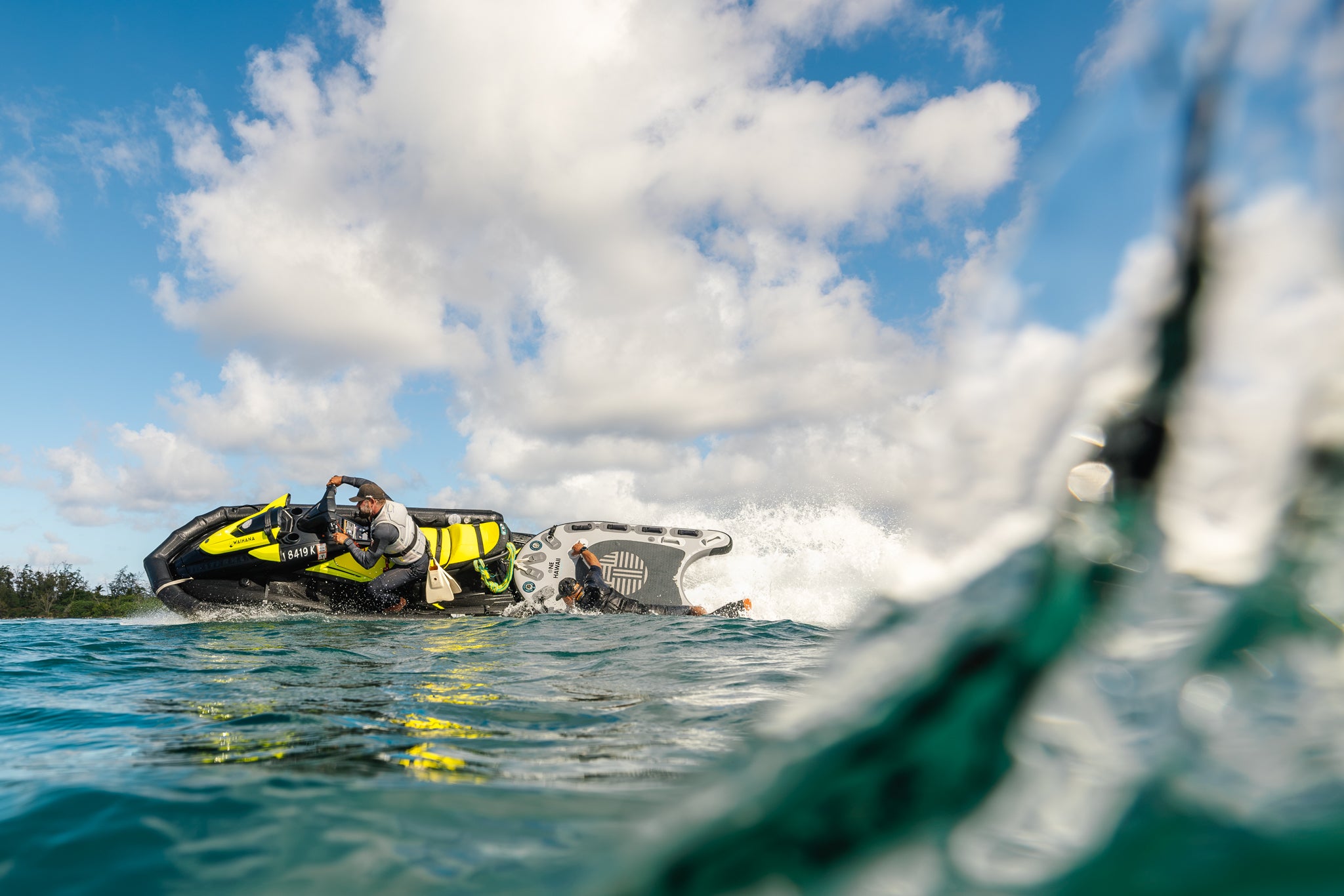 Two men wearing Waihana Tropicam Black Short Sleeve Spring Suits ride a Yamaha jet ski towing a rescue sled through choppy open ocean during waterman training, photographed from water level.