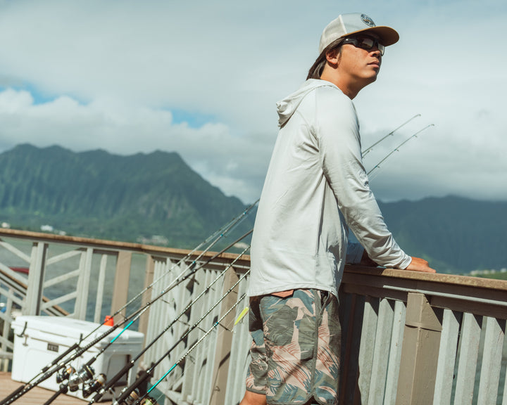 Man wearing a white hooded rashguard and Hawaiian Brushstroke Cruiser board shorts leans on a pier railing at Kaneohe Bay, Hawaii, with fishing rods and dramatic green mountains in the background.