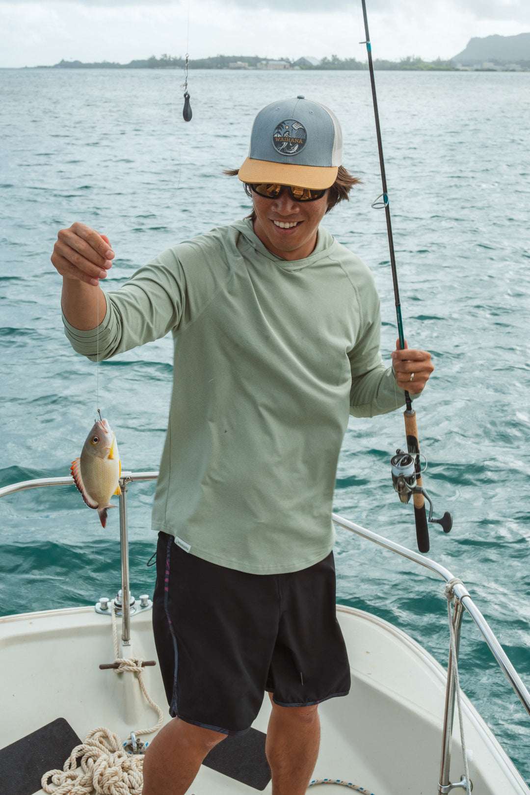 Smiling man in a Waihana snapback hat and sage green hooded long-sleeve rashguard holds a small fish and fishing rod on the bow of a boat in Kaneohe Bay, Hawaii.