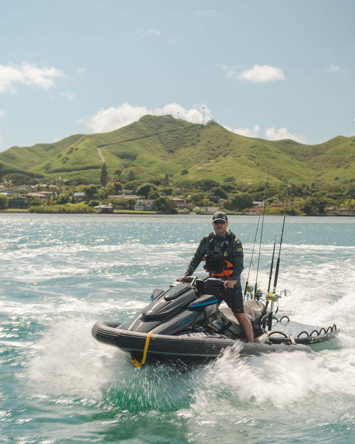 A man in a camo rashguard and hat rides a heavily rigged fishing jet ski across the turquoise waters of Kaneohe Bay, Hawaii, with green volcanic hills rising behind him.