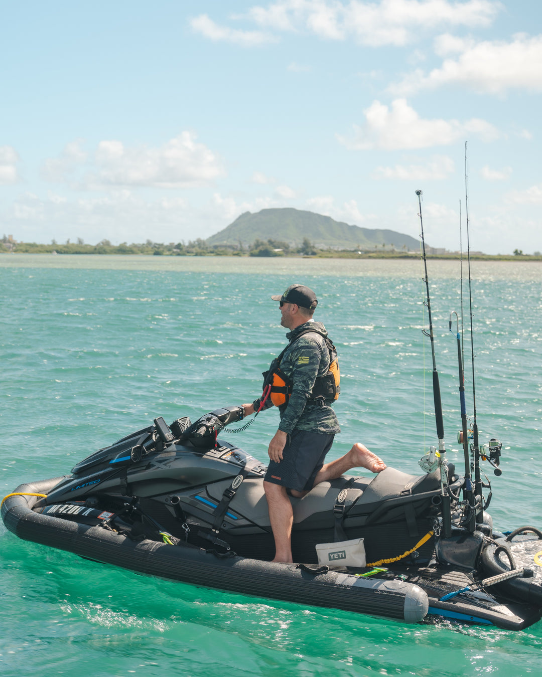 A man wearing a Waihana Tropicam Hydro Hat, camo long-sleeve rashguard, and Tropicam W1 Shorts stands on a fully rigged jet ski with fishing rods and a YETI cooler on the turquoise waters of Kaneohe Bay, Hawaii, with a mountain visible in the background.