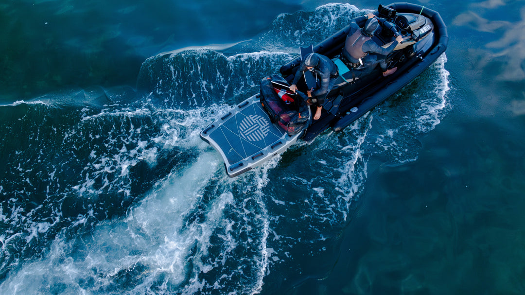 Aerial view of two wetsuit-clad spearfishers aboard a rigid inflatable boat speeding across deep blue ocean water, with spearfishing gear and a Waihana-branded deck mat visible.