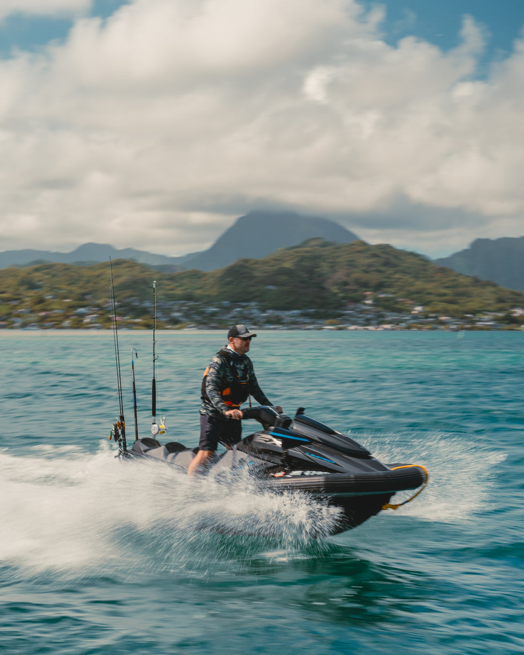 A man in a camo rashguard and cap stands on a black fishing jet ski with multiple rod holders, speeding across the turquoise waters of Kaneohe Bay with lush Hawaiian mountains in the background.