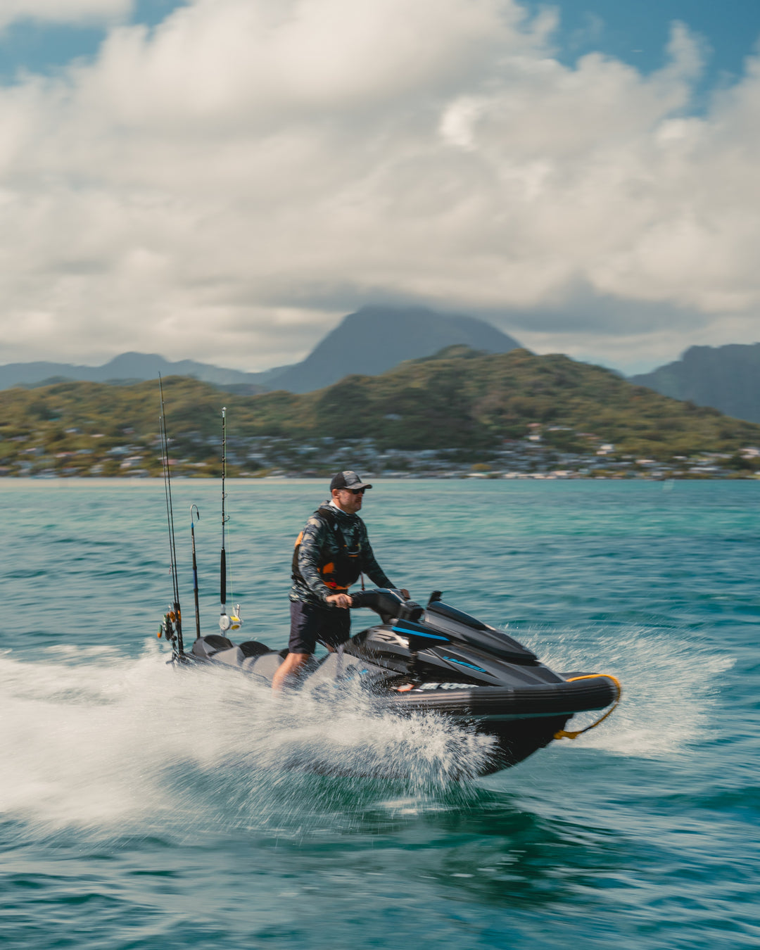 A man in a camo rashguard and cap stands on a black fishing jet ski with multiple rod holders, speeding across the turquoise waters of Kaneohe Bay with lush Hawaiian mountains in the background.