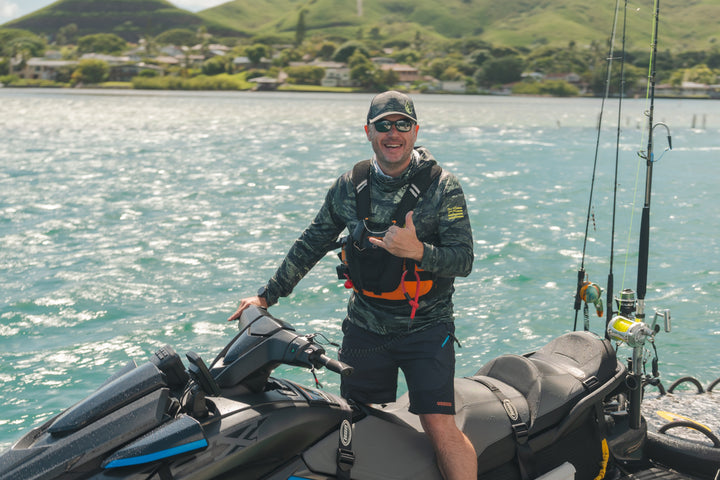 Smiling man in Waihana Tropicam camo sun shirt, hat, and board shorts giving a shaka sign next to a fishing jet ski on Kaneohe Bay with lush green hills in the background.