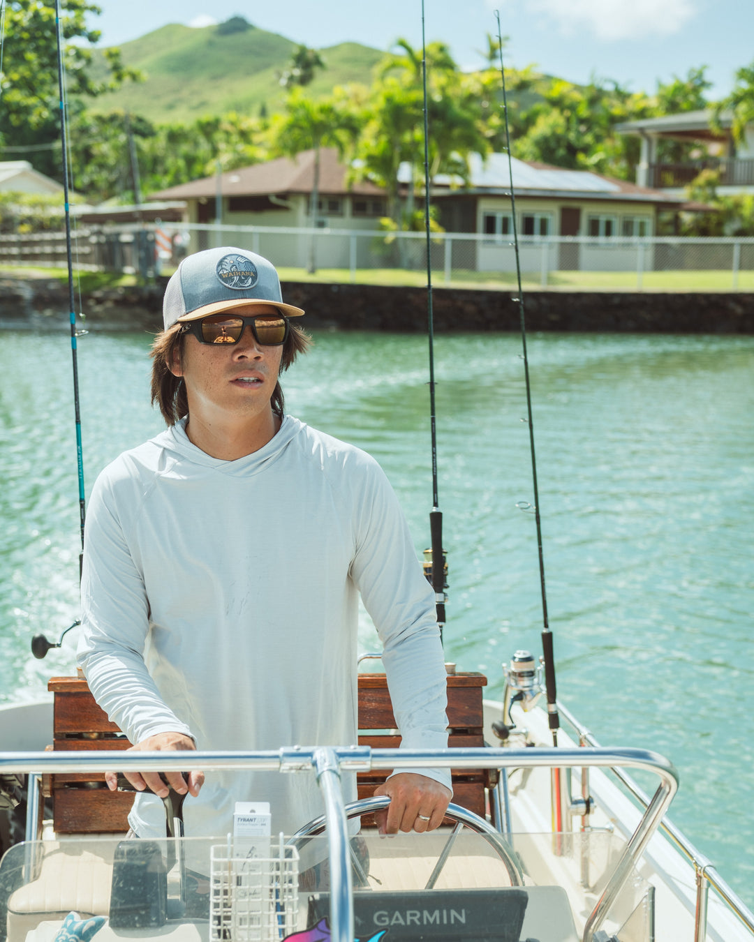 Man wearing a Waihana Lagoon Diver Trucker hat and white hooded long sleeve sun shirt steering a fishing boat in Kaneohe Bay, Hawaii, with green mountains and palm trees in the background.