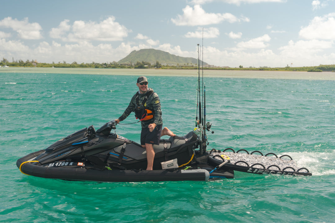 A smiling man wearing a Waihana Tropicam sun shirt, shorts, and hat stands on a heavily rigged jet ski with fishing rods and a side platform on the turquoise waters of Kaneohe Bay, Hawaii, with a green volcanic mountain in the background.