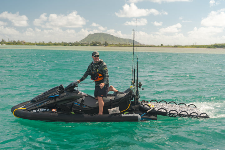 A smiling man wearing a Waihana Tropicam sun shirt, shorts, and hat stands on a heavily rigged jet ski with fishing rods and a side platform on the turquoise waters of Kaneohe Bay, Hawaii, with a green volcanic mountain in the background.