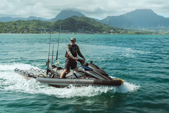 Man standing on a fishing jet ski loaded with rod holders cruising across turquoise Kaneohe Bay waters wearing a Waihana Tropicam camo hat and board shorts, with lush Hawaiian mountains in the background.