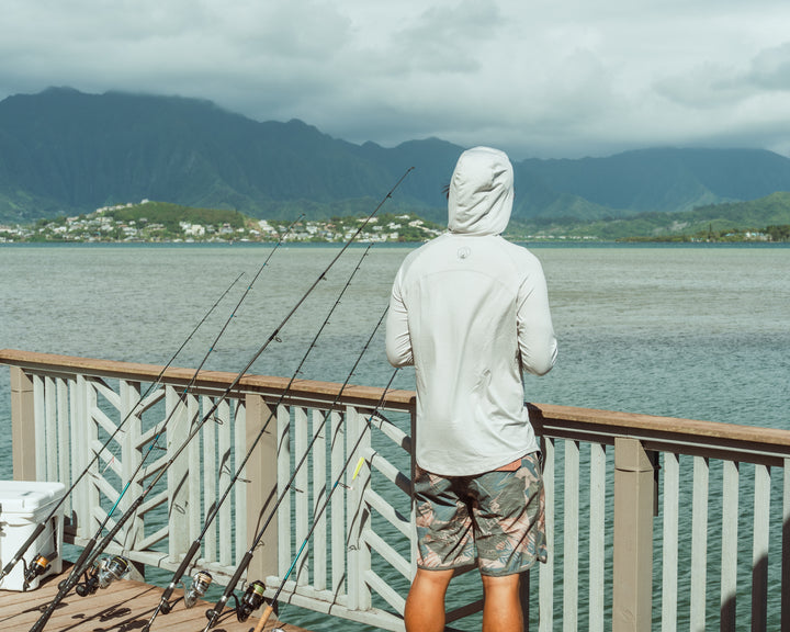Man wearing a white hooded sun shirt and Waihana Hawaiian Brushstroke Cruiser Shorts stands on a wooden pier at Kaneohe Bay, surrounded by fishing rods with lush Hawaiian mountains in the background.