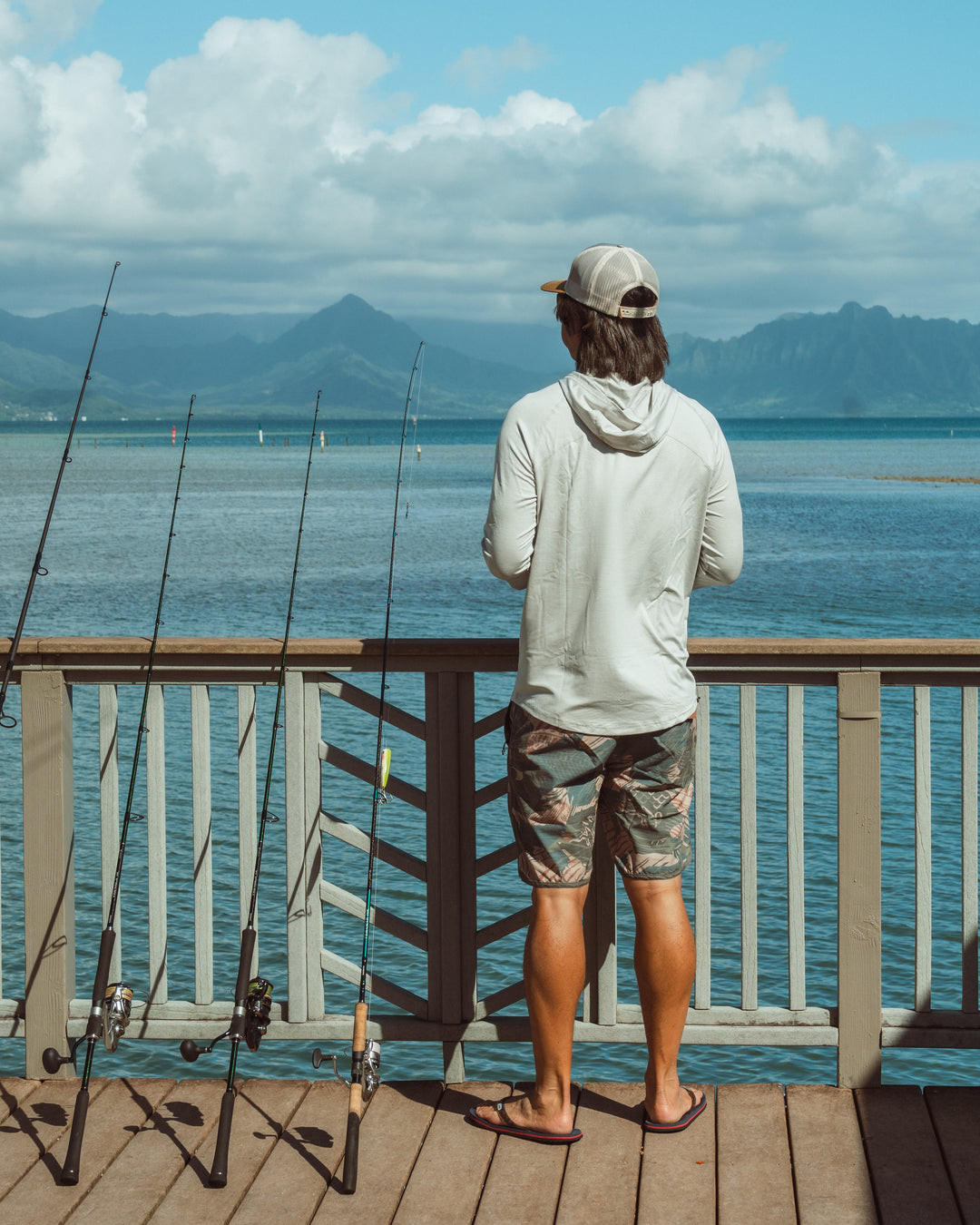 A man in a white hooded rashguard, camo shorts, and a snapback cap stands on a wooden pier at Kaneohe Bay, Hawaii, looking out at the water with several fishing rods leaning against the railing and the Ko&