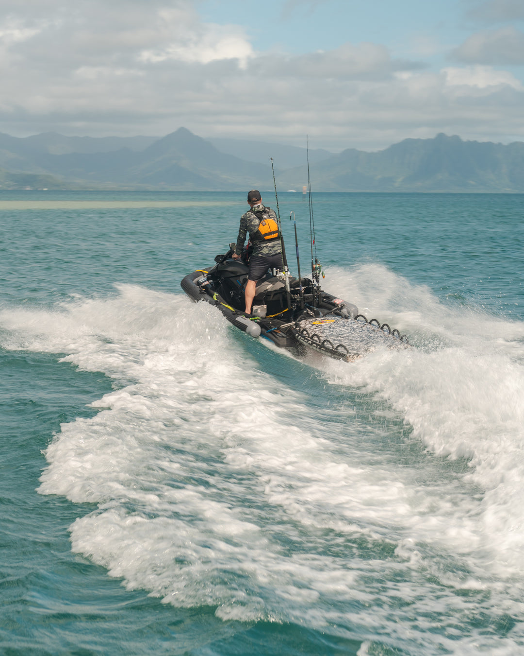 A man wearing a camo long-sleeve rashguard, orange life vest, and hat stands on a gear-loaded jet ski with fishing rods, speeding across the turquoise waters of Kaneohe Bay with misty green mountains in the background.
