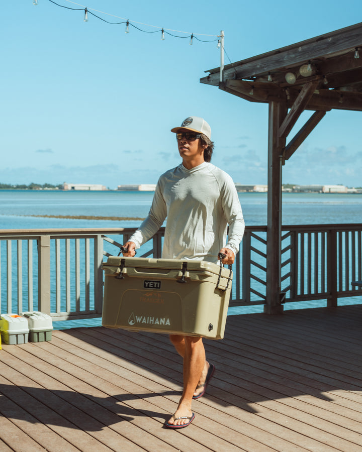A man in a white long-sleeve rashguard and trucker hat carries a large Waihana-branded Yeti cooler across a wooden waterfront deck with calm ocean waters visible behind him.
