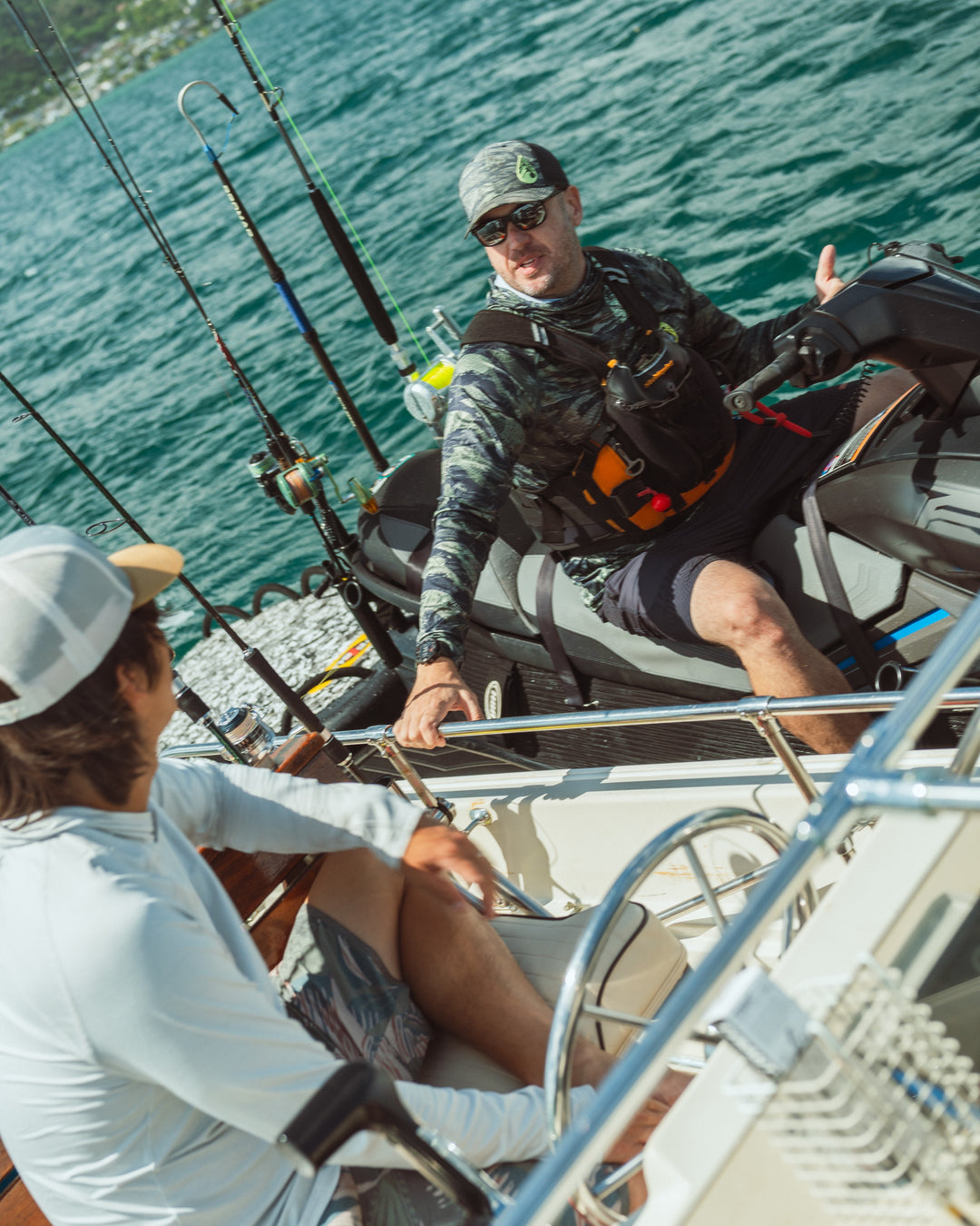 A man on a jet ski wearing a Waihana Tropicam camo hat, camo rashguard, and orange life vest talks with another person on a nearby boat surrounded by fishing rods on Kaneohe Bay.