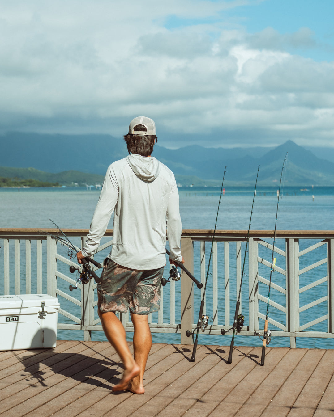 A man seen from behind on a wooden pier at Kaneohe Bay, holding multiple fishing rods, wearing a white long-sleeve hooded rashguard and camo shorts, with a Yeti cooler to his left and lush Hawaiian mountains across the water.