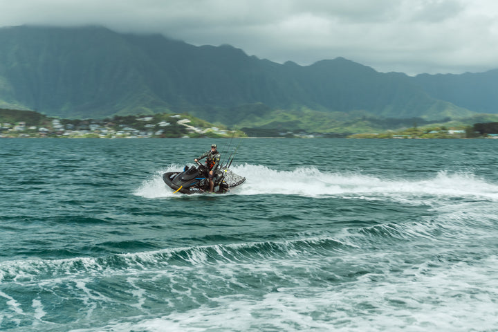A man in a camouflage wetsuit rides a black jet ski loaded with fishing rods across the choppy turquoise waters of Kaneohe Bay, with lush green Ko&