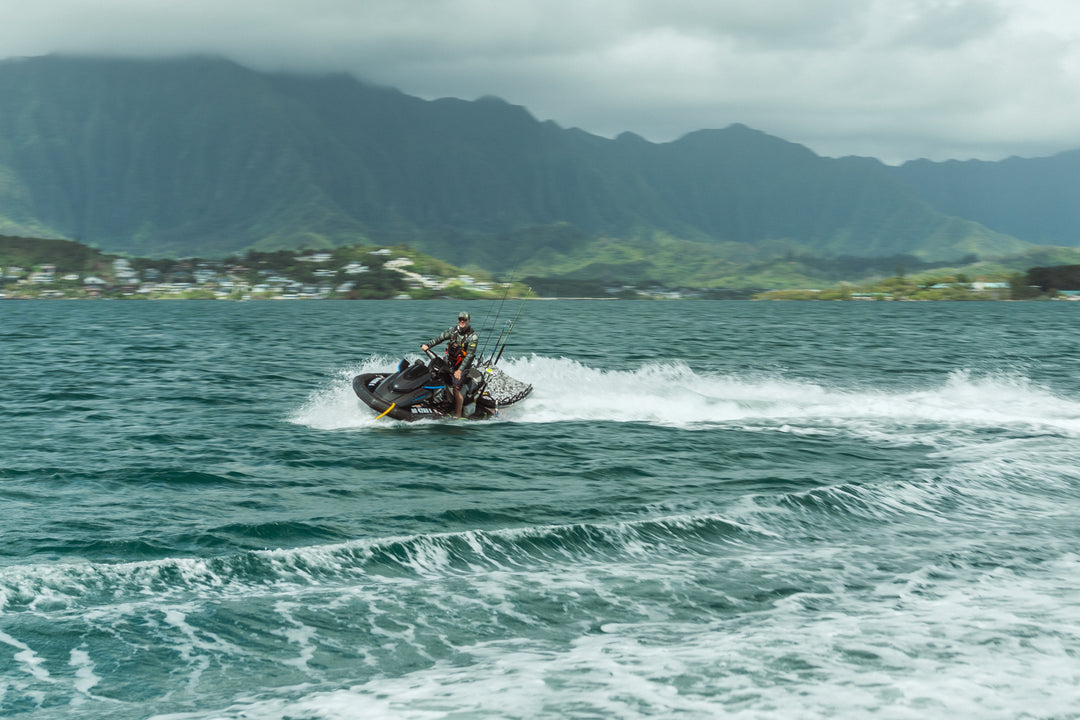 A man in a camouflage wetsuit rides a black jet ski loaded with fishing rods across the choppy turquoise waters of Kaneohe Bay, with lush green Ko&